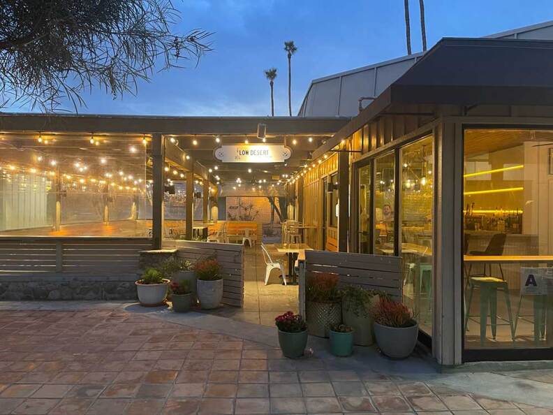 An exterior shot of an entryway to a restaurant with a brick walkway leading to an outdoor patio enclosed with lights, tables, and chairs. Next to the deck is the restaurant's interior, which can be seen by the full-sized windows next to the patio space.