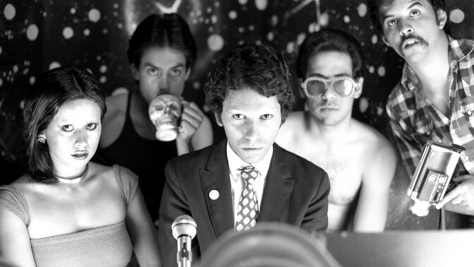 Five people pose closely together, one drinking from a skull-shaped mug and another wearing heart-shaped sunglasses, against a starry backdrop.