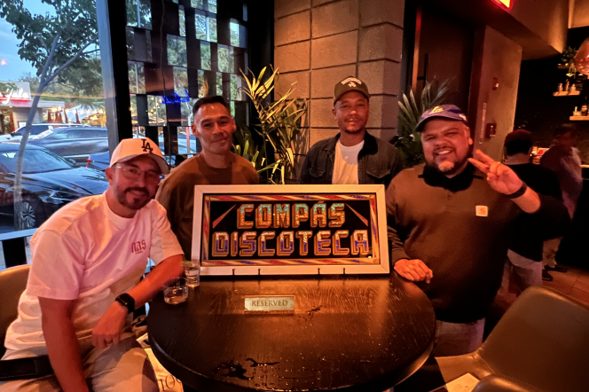 Four men pose at a table around a sign that says "Compás Discoteca". The one on the far left wears a white "LA" Dodgers hat. The one to the right of him wears a brown shirt. The one to the right of him wears a hat, blue jacket, and white shirt. The one one on the far right wears a blue hat and a "Carhartt" jacket.