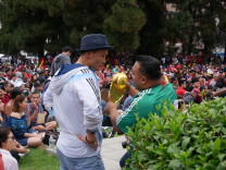 Two men with dark skin stand in front of a crowd talking to each other. One is wearing a light blue jacket and hat with his hands at his side. The other is wearing a green shirt with white stripes on the shoulders while holding a gold trophy. 