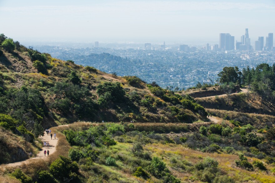 A trail populated by hikers winds through a hilly landscape. A skyline is in the distance.