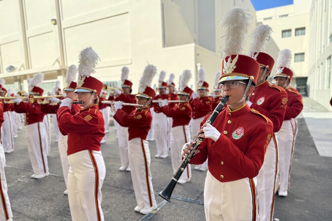 Rows of marching band members in red and white uniforms play instruments outside a white building.