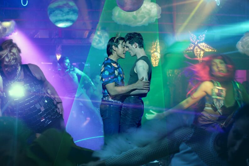 A movie poster for 'glitter & doom' featuring two men holding each other close under strobe lights.