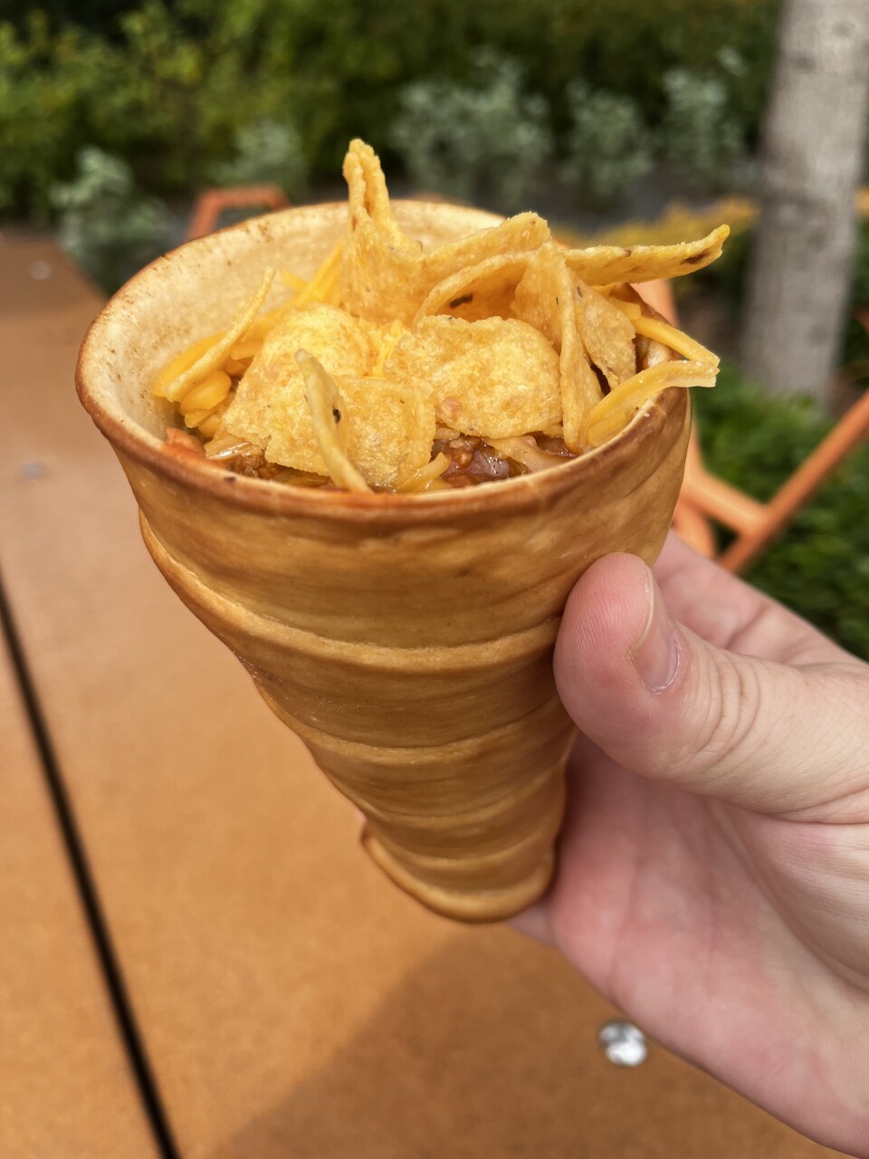 A light-skinned hand holds a cone made of baked dough filled with chili and topped with yellow corn chips.