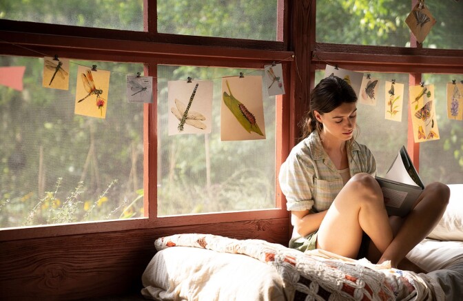 Daisy Edgar Jones sits on a twin-sized bed pushed up against two large windows with wooden frames. She sits crossed legged, her legs propping up a book she is perusing. Twine string is stretched horizontally over the windows, with clips holding up various drawings of swamp life i.e. cattails and dragonflies. Outside the windows, trees and greenery can be seen.