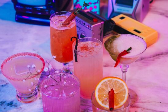 Six colorful cocktails are displayed on a marble table under blue and pink lights. Behind them are examples of nostalgic technology, like a Talkboy, a vintage portable TV and a Gameboy.