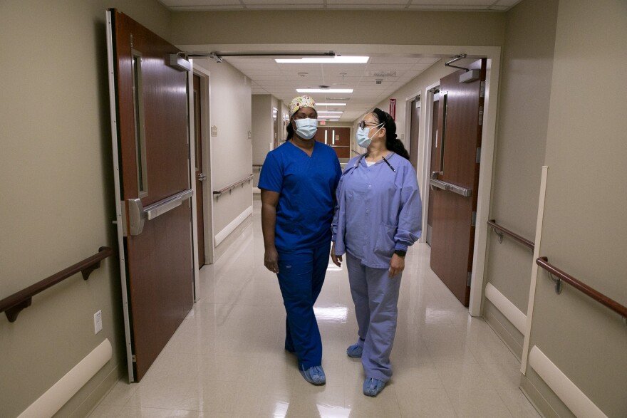 Two nurses, one in deep blue scrubs and the other wearing light blue, stand in a hospital corridor with masks on.