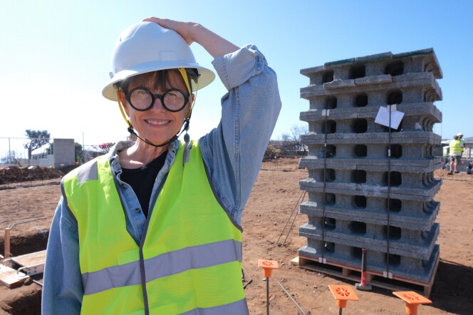 A woman with medium light skin tone, wearing a hard hat and fluorescent vest, stands in front of a stack of gray concrete blocks on a dirt lot in the Los Angeles County neighborhood of Sunset Mesa. 