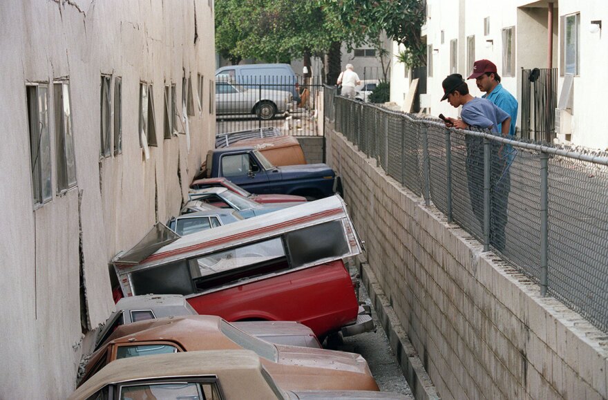 An apartment building collapsed, crushing cars. Two kids look at the destruction from a fence nearby.
