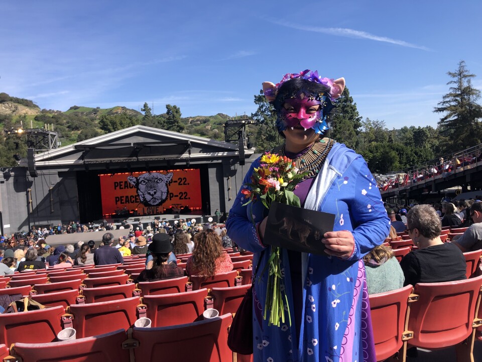A woman stands in an aisle at an outdoor theater wearing a pink and blue mask in the shape of a mountain lion. She's holding a bouquet of flowers.