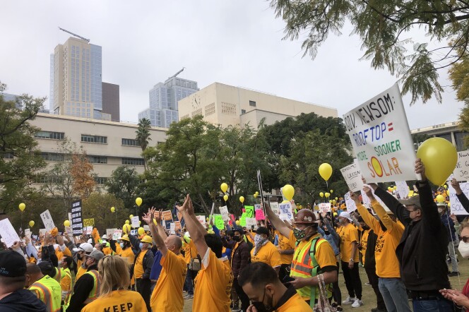 A crowd of people in yellow shirts with signs and yellow balloons cheer with hands up in a grass field in Grand Park in downtown L.A. as buildings loom in the background. 