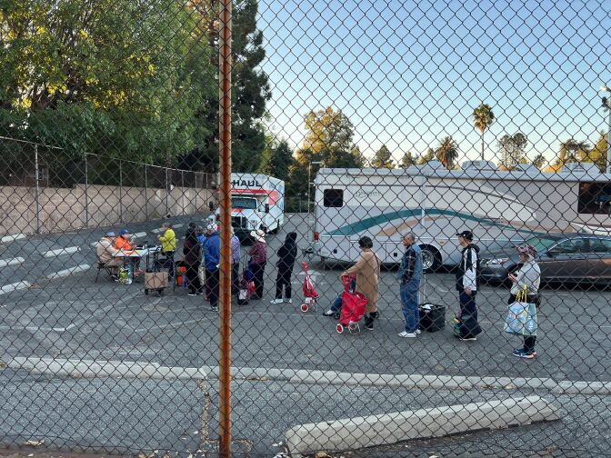 A group of people gathered in an outdoor parking lot, viewed through a chain-link fence.  