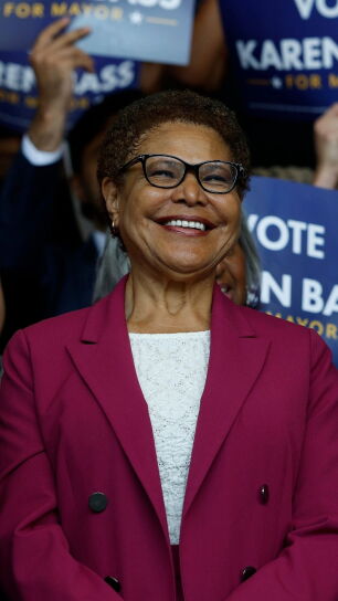 A smiling woman with short curly hair and glasses, wearing a magenta blazer over a white top, stands surrounded by supporters holding "Vote Karen Bass for Mayor" campaign signs.