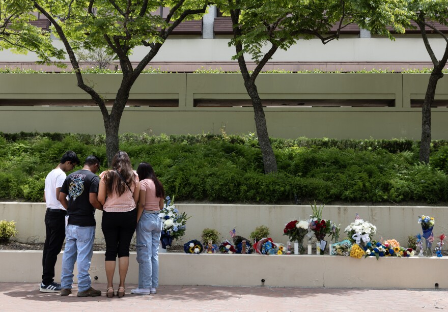A small group of people bow their heads in front of a makeshift memorial covered in flowers.