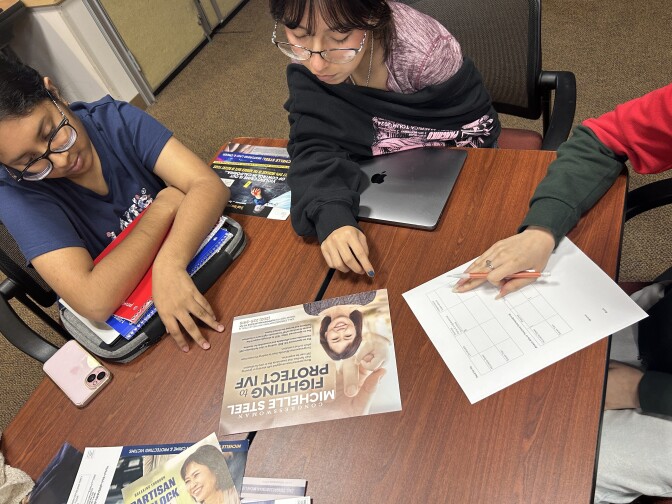 Students sit around a long folding table, all peering down at a political mailer that is on the table. One of the students has a white sheet of paper and is poised to write down group impressions about the campaign literature. 