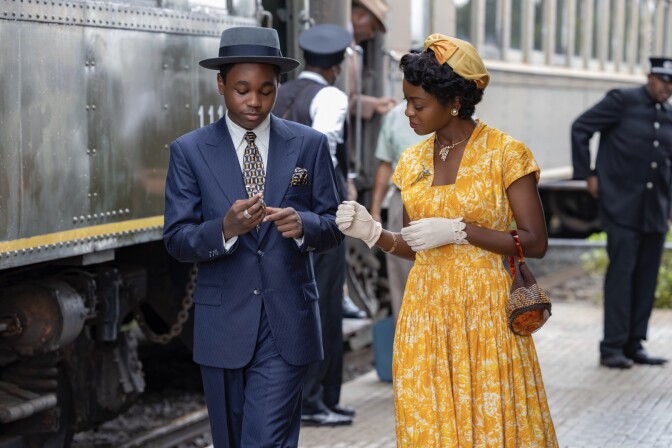 Jalyn Hall stands mid-stride on the left in a navy blue suit. He is looking down at his slightly clasped hands. His left hand has a prominent silver ring on the middle finger. On Hall's right is Danielle Deadwyler in a yellow dress with white florals. She wears a matching hat and white wrist gloves. Her right hand rests slightly below her chest as her left hand motions towards Hall's hand as if they are passing something between them.  She is also looking down at Hall's hands seemingly holding something of interest. Behind them to the left is a train car with people coming out of its doors. There is a conductor in the back right indicating that they are on at a train platform, possibly just arriving. 