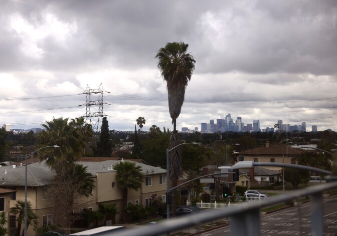 The skyline of Los Angeles in the background surrounded by heavy gray clouds. In the foreground a neighborhood as seen from above. 