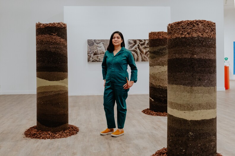 A woman with medium-light skin tone and medium-length dark hair stands among three 6-foot tall cylinders made of soil, clay, zeolites, and mulch.