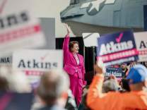 A woman with light skin, close cropped red hair and wearing bright pink pants and a bright pink blazer over a lighter pink blouse raises her hand in the air as she is surrounded by dozens of people holding Atkins for Governor placards