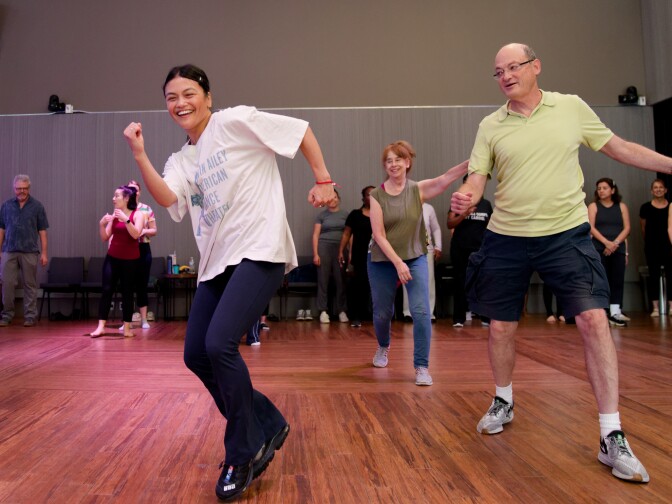 A woman in black leggings and a matching long-sleeved crop top stands barefoot on a wooden studio floor. She has her eyes closed and palms open in a peaceful, grounded pose, while other participants in athletic wear stand behind her.