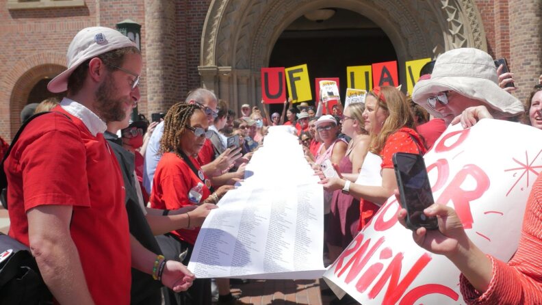 About three dozen people, mostly clad in red tops, hold up a long scroll filled with signatures as they rally in front of a brick campus building.