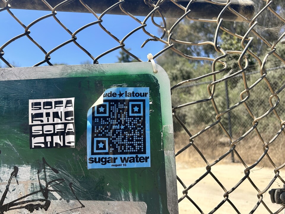 A blue sticker with a qr code and the phrases "Maude Latour," "Sugar Water" and "August 16," plastered on the corner of a green sign on a chain-link fence