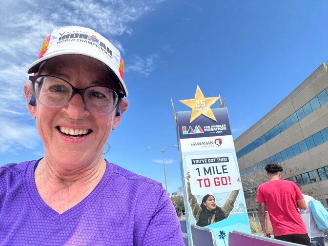 A woman in a purple running shirt, hat and glasses standing in front of a sign that says, "1 Mile To Go!"