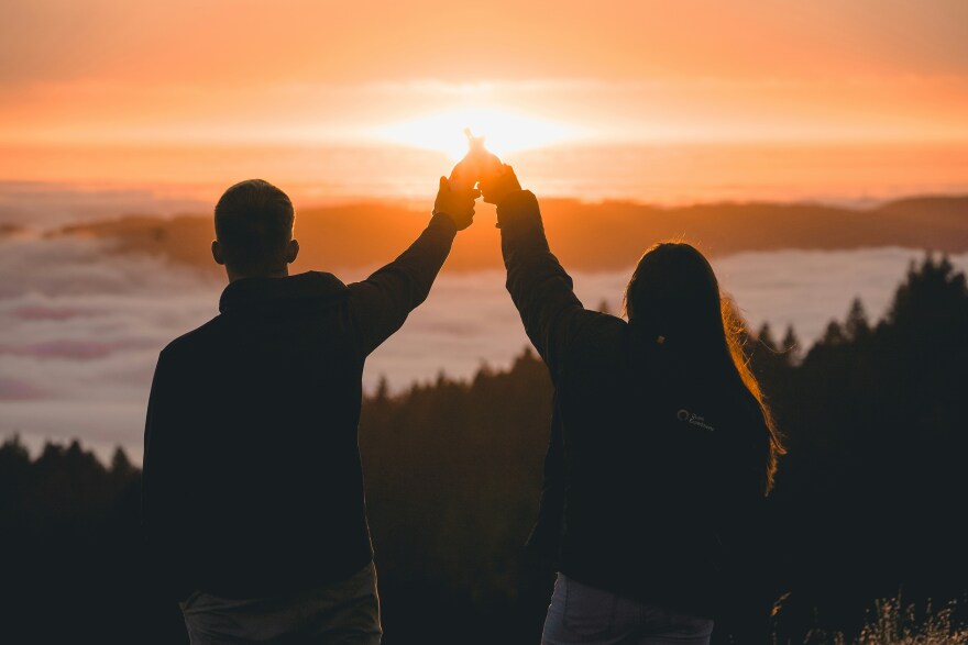At sunset, two people stand in silhouette with their backs to the camera, their arms raised high in a "cheers" gesture, they hold two bottles that touch in the air. In the background is a forest view below them with tall trees peaking out between clouds along the horizon.