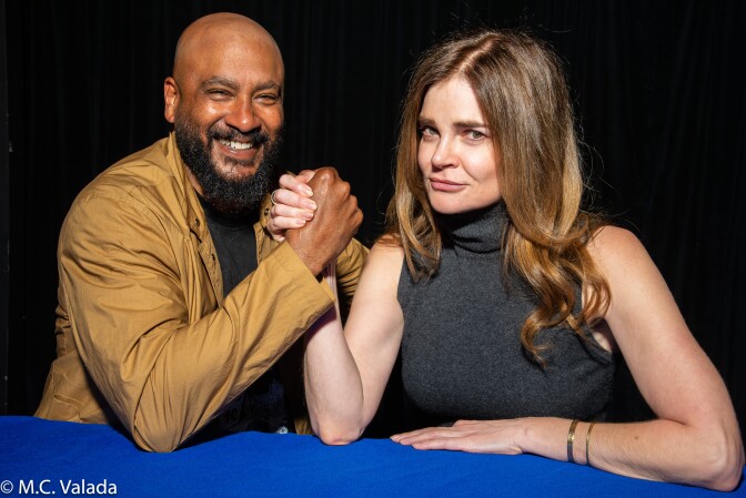 Cirroc Lofton, a Black man with bald head and beard and Betsy Brandt, a white woman with long brown hair, grasp hands and smile at camera