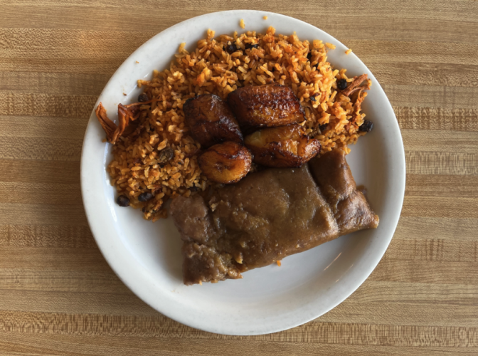 An overhead shot of a white plate on a wood table featuring arroz con gandules, caramelized sweet plantains, pasteles, and pulled pork pernil.