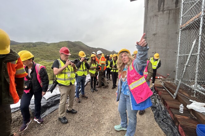 A woman with long blonde hair, wearing a pink high-visibility vest and a yellow hard hat, is pointing towards the camera with her arm outstretched. She's standing in an outdoor construction zone, surrounded by a group of men and women wearing similar yellow hard hats and neon yellow or orange vests.