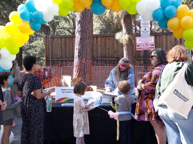 kids in line for books. ballons at top of frame. a sign reads 'books and bags for children'