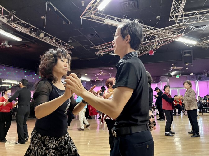 An Asian American woman in black holds raised hands with an Asian American man, also wearing black, as she dances.
