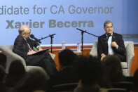 Two men sit on a stage in upholstered chairs with mics. The backs of students can be seen.  