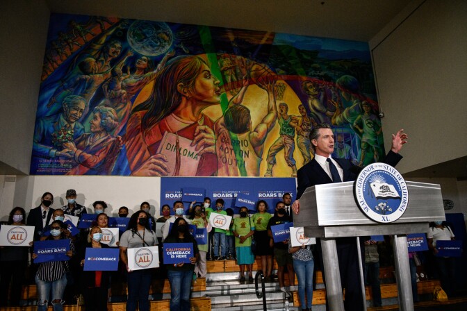 Gov. Newsom in a suit and tie stands at a lectern in front of a group carrying signs supporting him.