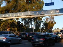 Cars are backed up across four lanes heading into Dodger Stadium, under a Dodger Stadium banner that notes it's at GATE A, with a street sign reading STADIUM above, and traffic lights to the left of that.