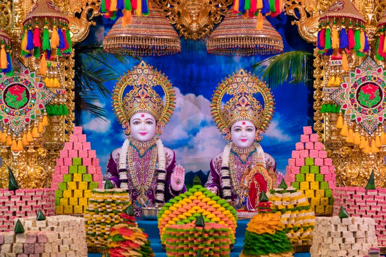 A shrine is made up of two characters. Both have their right hand up showing a small symbol on their palm and are wearing a golden headdress. Surrounding them are towers of different types of sweets that are multicolored. There are also large tapestries on both sides made up of gold as well as different colored tassels.