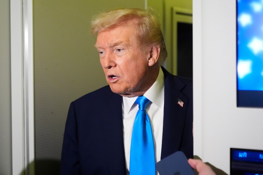 Donald Trump stands in a doorway. He wears a dark suit and bright blue tie.