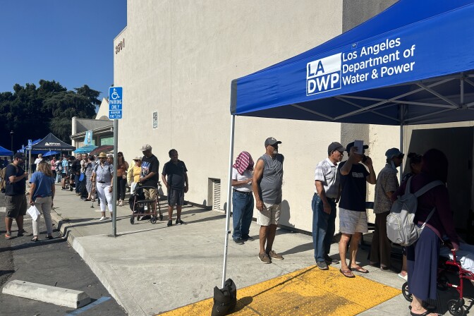 People wait in line under the sun beside an off white building. There's a shade ten with blue awning and white lettering "LADWP"