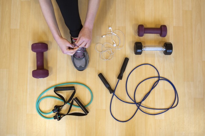 A person ties a sneaker on a wood floor with exercise equipment including dumb bells and a jump rope.