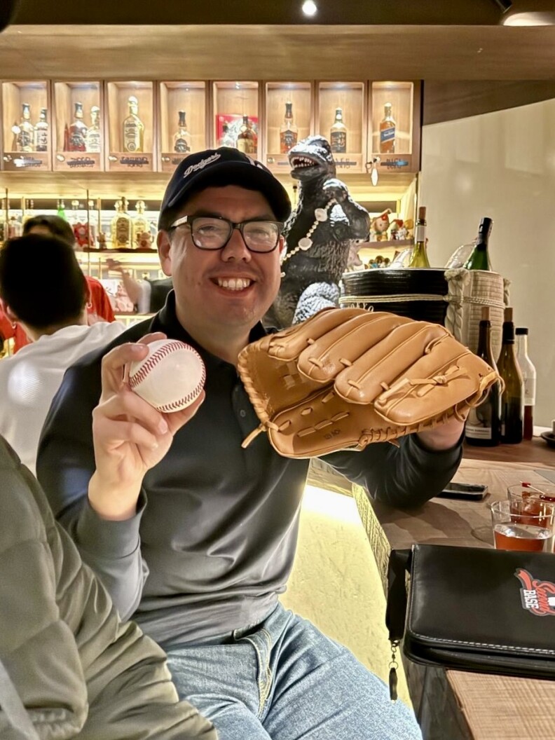 A 30-something man wearing glasses and a baseball cap poses in a bar holding a baseball glove.