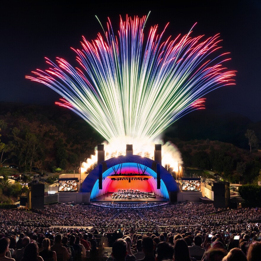 Fireworks shoot out from behind a stage during an outdoor show at night with a large crowd watching.