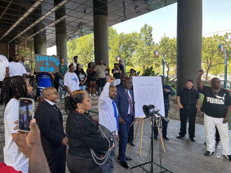 A group of people gather near the steps to a building with round columns. Some have their firsts raised. One holds a sign that says: Justice for Rob. Most have dark-tone skin.