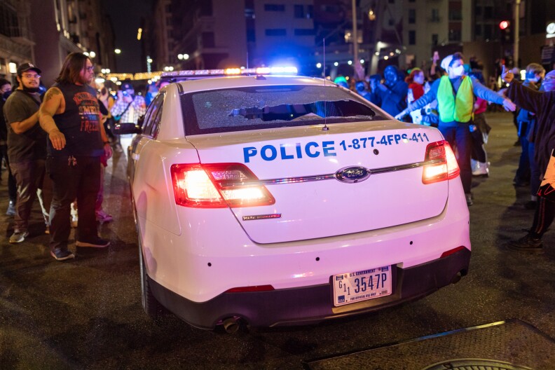 A group of people crowd around a police car with a back broken glass window.