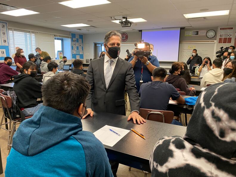 A man in a suit and facemask stands at a desk in a classroom, talking to students. Media members with cameras are in the background.