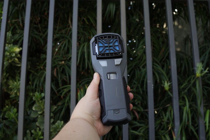 A hand with a light skin tone holds the rectangular gray device into the air front of plants and metal bars. The device has a black gill on the top end with a blue mat underneath by the buttons.