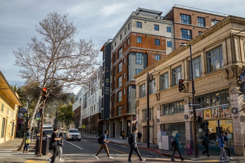 A view of a city street while people walk across it by a stoplight. There are multiple buildings and trees in the view.