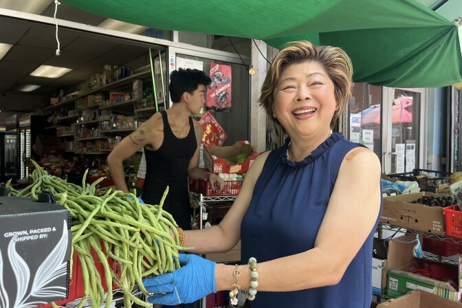 An Asian woman stands at the entrance of a small market under a green tarp, smiling and holding a bunch of long green beans. Behind her, a younger Asian man stands amid stacked produce boxes, and shelves of groceries line the back.