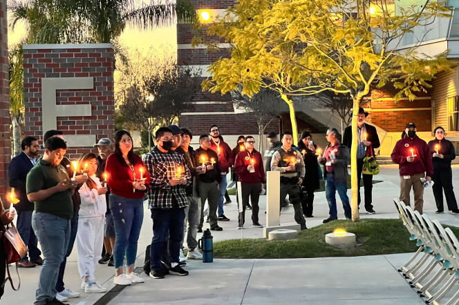 A group gathers around a tree in a campus quad holding candles. The trees are uplit as it is evening and a brick column with a large E on it 