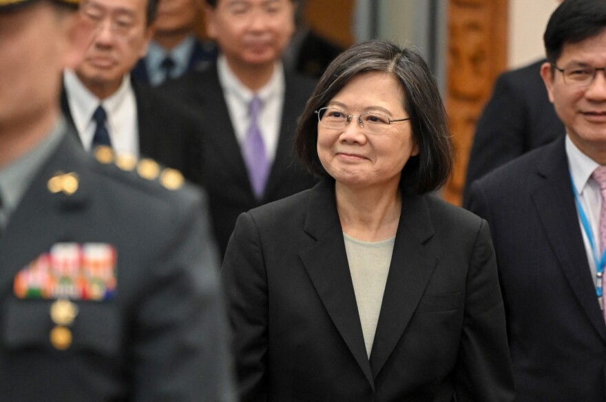 An Asian woman who appears to be in her mid-60s wears a black jacket and a light shirt as she arrives at an airport gate. She is looking off to the left with a courteous grin as she acknowledges people around her.